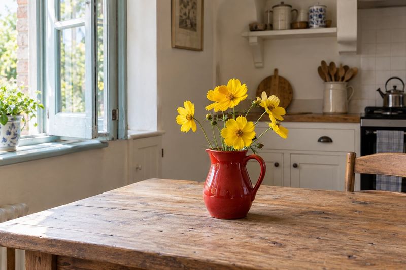 Yellow flowers in a red jug on a wooden kitchen table, with the room and window around them.